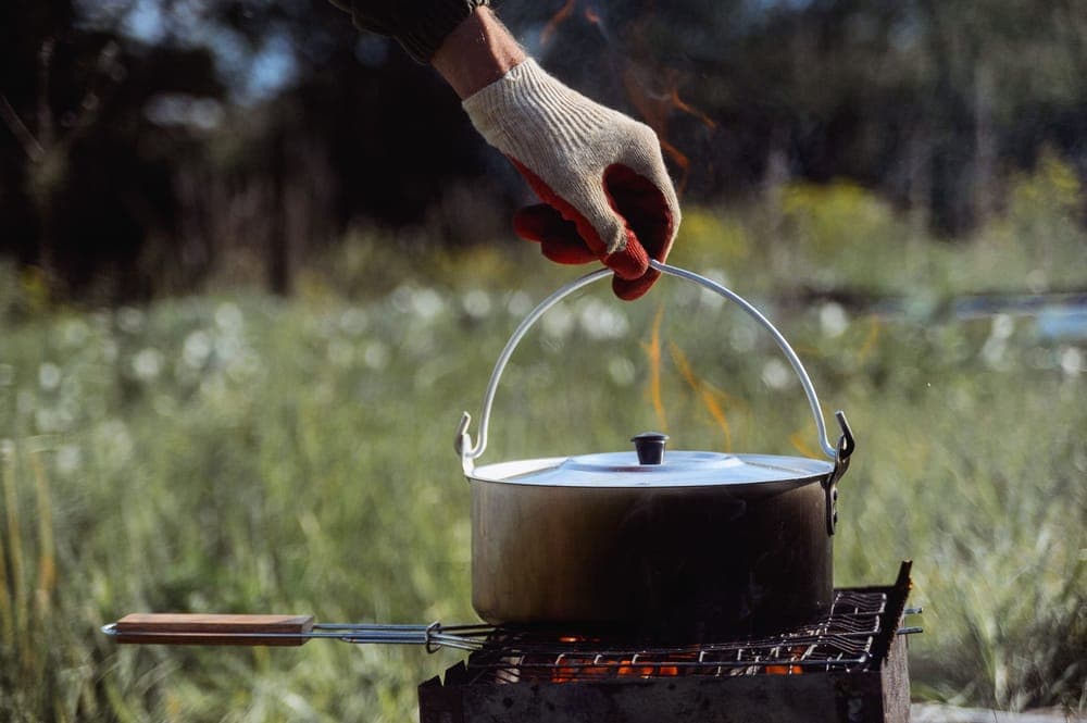A gloved hand holding a pot over a campfire grill