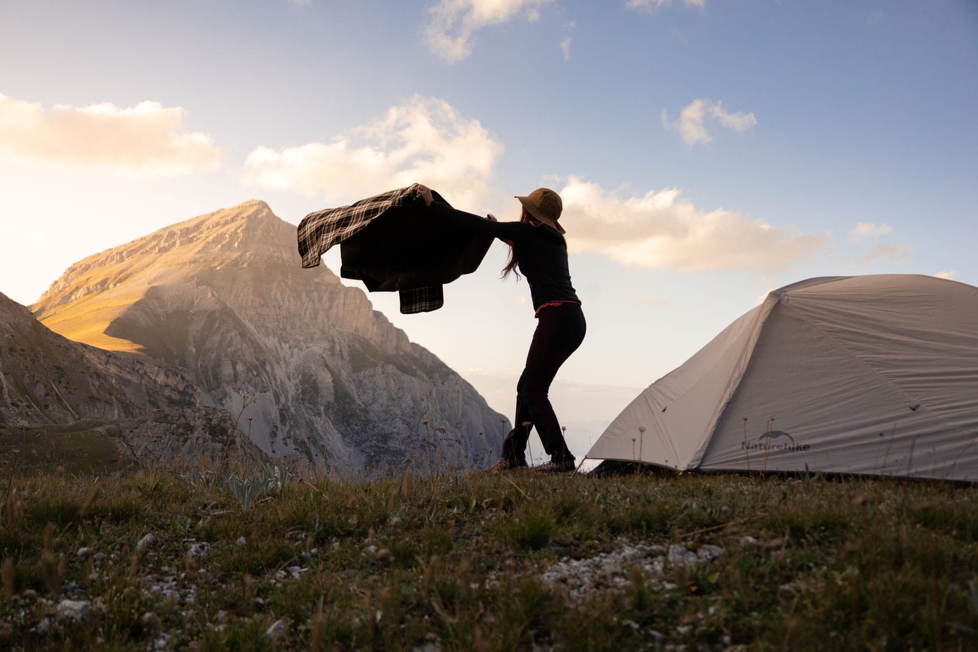 A hiker shaking out a blanket beside a tent on a mountain meadow at sunrise